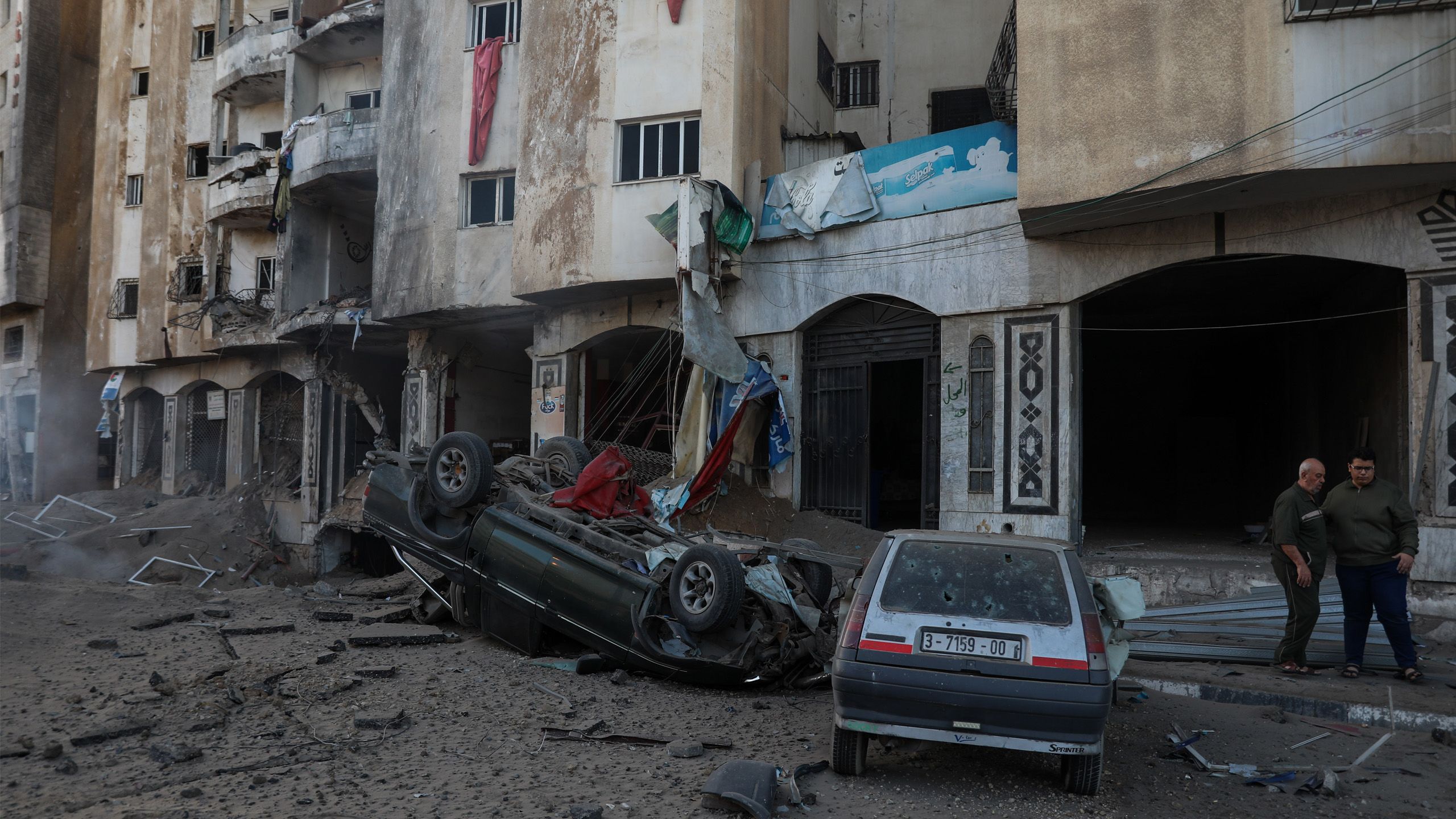 A bombed building in Gaza. The walls are torn out, and debris are strewn everywhere. In front of the building is a car flipped up side down.