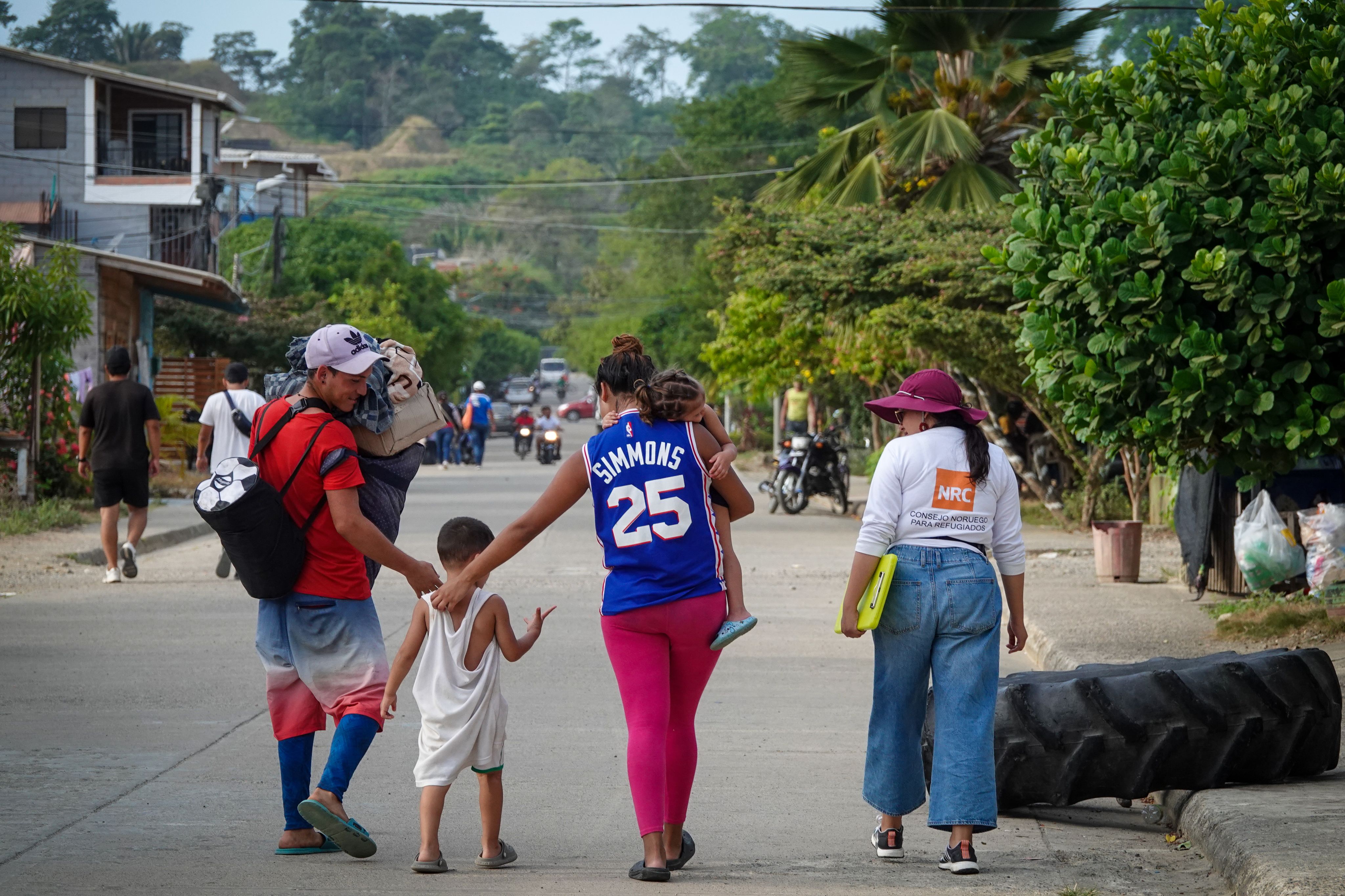 Alexander walking with his wife and children in a street in Necoclí. An NRC employee walks to the right of them. They are surrounded by trees and other people walking.
