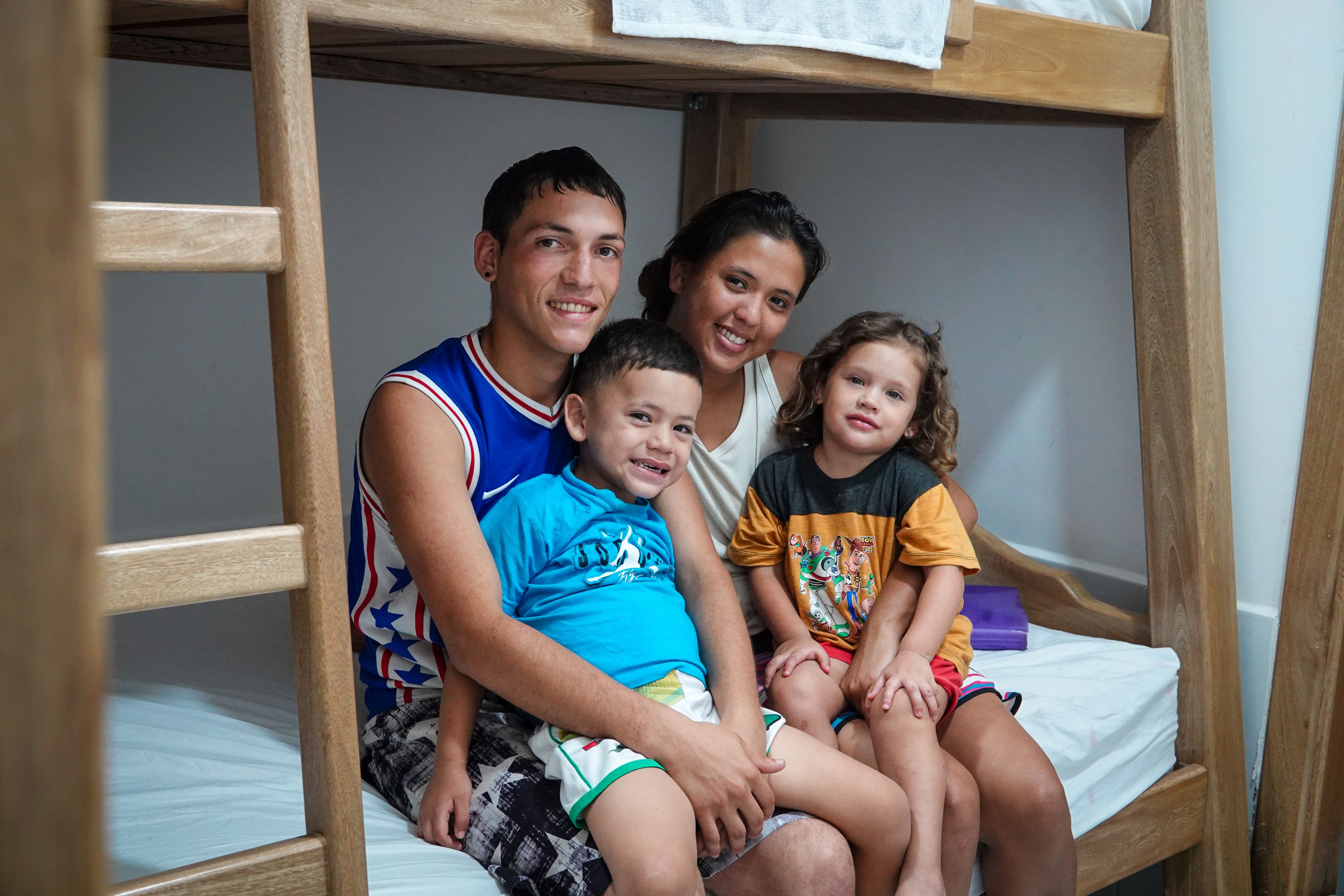 Family of four (father, mother, son and daughter) sitting on the lower bunk of a bunk bed smiling at the camera.
